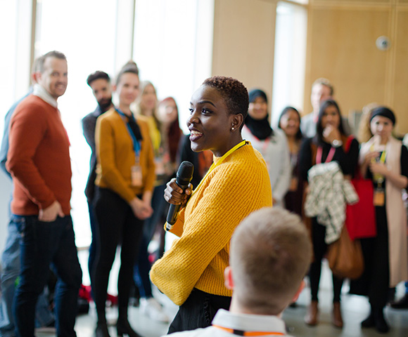 woman speaking to an engaged audience wearing a yellow sweater group of five people in background smiling and listening intently